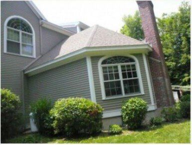 With LeafLock — Gutter Covers in Portsmouth, NH A tan, two-story house with a brick chimney, grey siding, and arched windows, surrounded by green landscaping.
