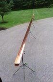 A long, wooden V-shaped chute mounted on two tripod stands, positioned outdoors on a paved surface.