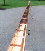 A long copper gutter section rests on metal stands on a paved driveway near a grassy lawn.