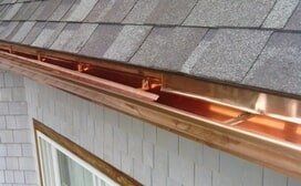 A close-up view of copper gutters installed along the edge of a gray shingled roof above a wood-trimmed window.