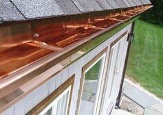 A high-angle view of shiny, newly installed copper gutters attached to the edge of a roof above a home with white siding.