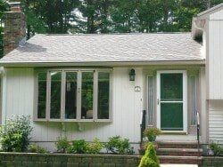 A single-story home with light siding, a bay window, and a green front door under a small porch with brick stairs.