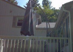 A closed, dark-colored hanging patio umbrella stands on a wooden deck next to a house under a clear, sunny sky.