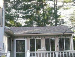 A single-story home addition with a grey shingled roof, glass sliding doors, and a white deck railing, set against trees.