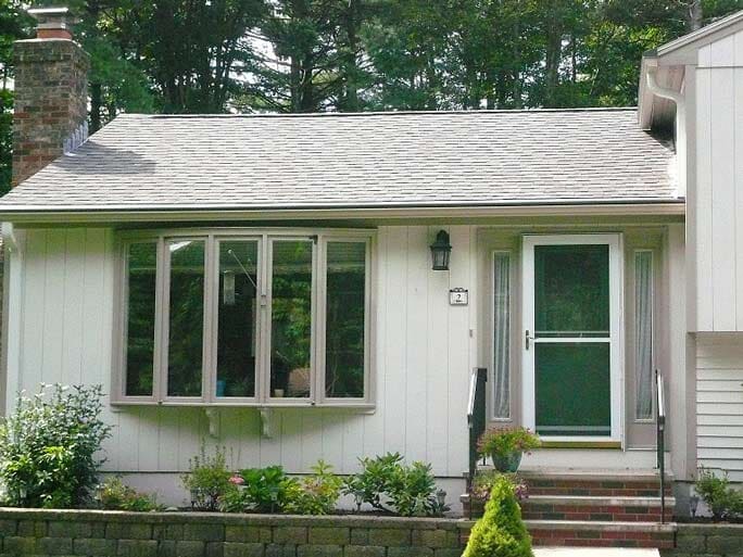 A light-colored house exterior with a large bow window, a storm door with steps, and a stone chimney.
