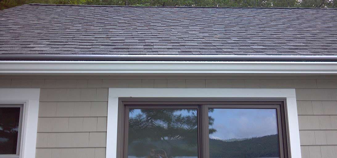 A tan house exterior with white window trim, a horizontal window, and a grey shingled roof under a gutter.