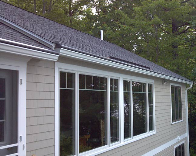 A side view of a house with light tan shingle siding, white trim windows, and a dark grey shingled roof.