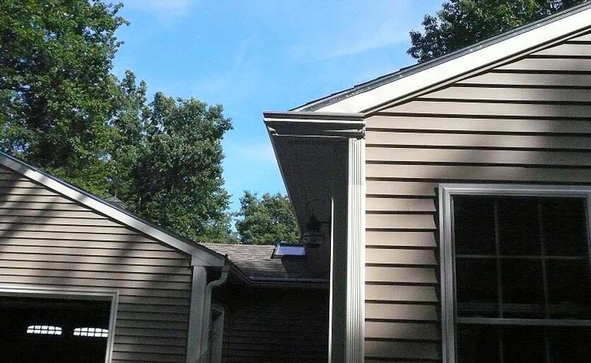 Exterior view of a beige-sided house with a dark roof, a white gutter system, and a window, framed by green trees.