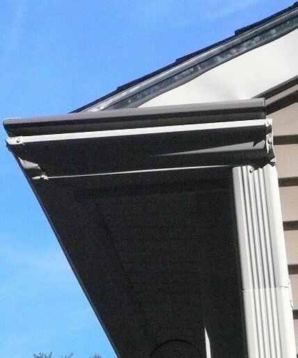 The corner of a building's roof with a dark brown gutter and a white, fluted downspout against a clear blue sky.