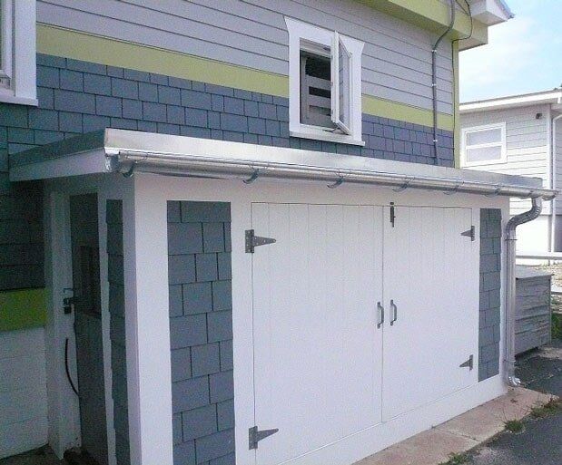 A small white storage shed with grey accents attached to the side of a larger house with matching siding and a metal roof.