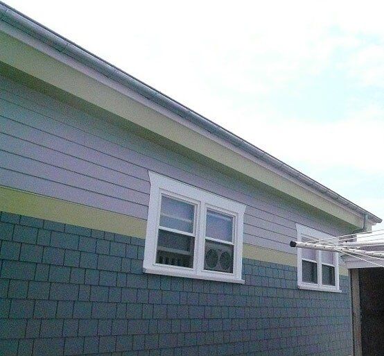 Exterior view of a house wall with green square shingles, horizontal lavender siding, and two white-framed windows.