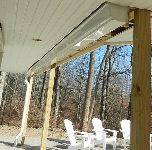 A white electric patio heater mounted to the ceiling of a covered porch, overlooking a wooded area with white chairs.