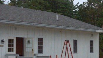 A white building with vertical siding, a grey shingled roof, a ladder, and two doorways, framed by trees.