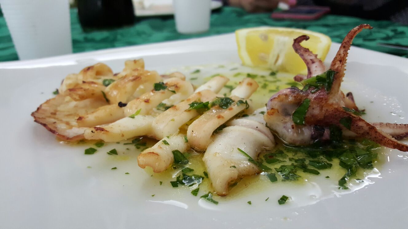 Frutti di mare grigliati serviti su un piatto bianco con salsa alle erbe aromatiche e spicchi di limone.