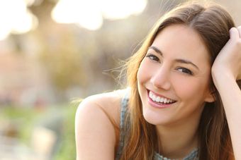 A smiling person with long brown hair leaning their head on one hand, captured in soft, bright outdoor light.