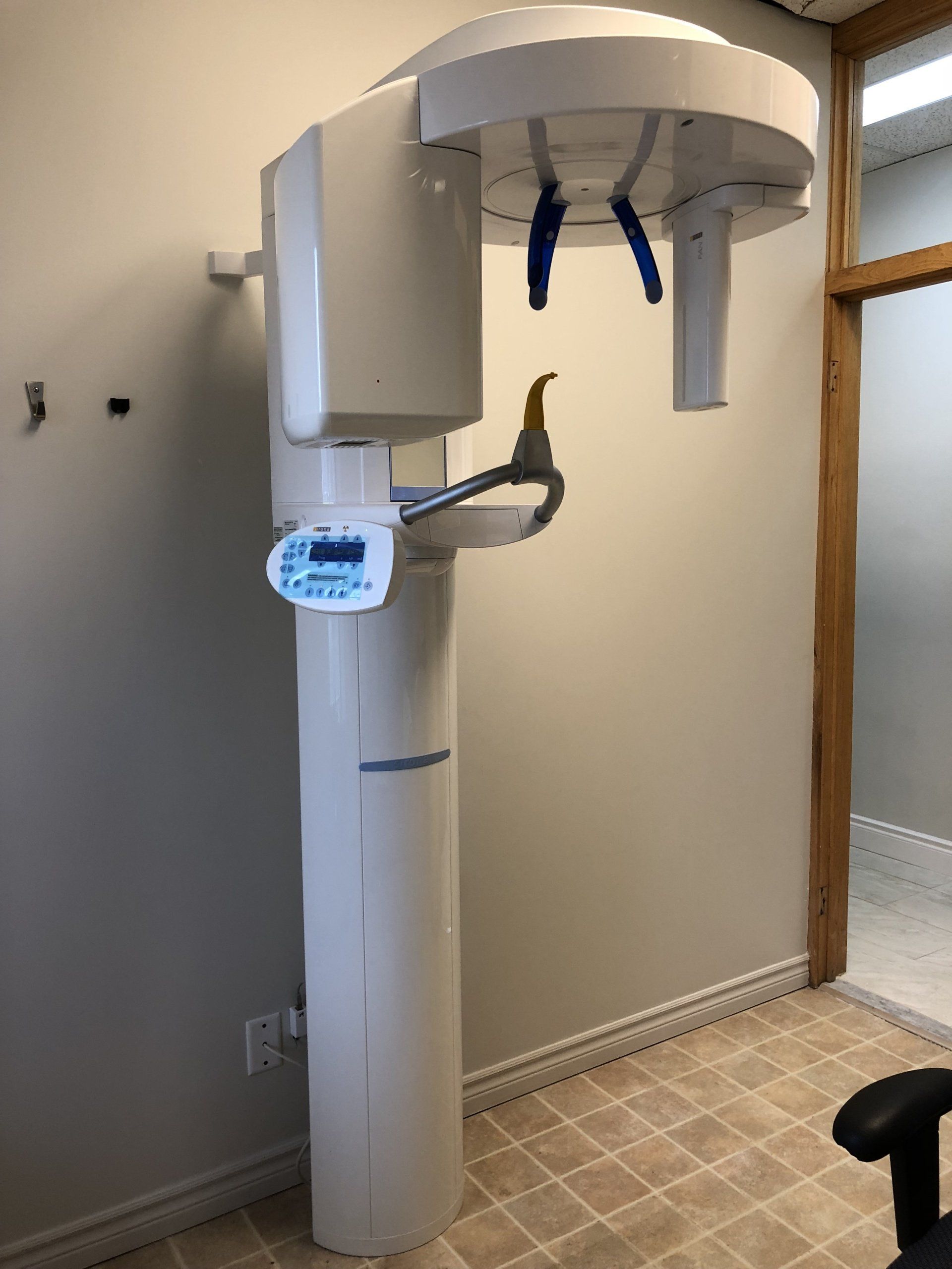 A wall-mounted white dental panoramic X-ray machine standing in a clinical office with tiled flooring.