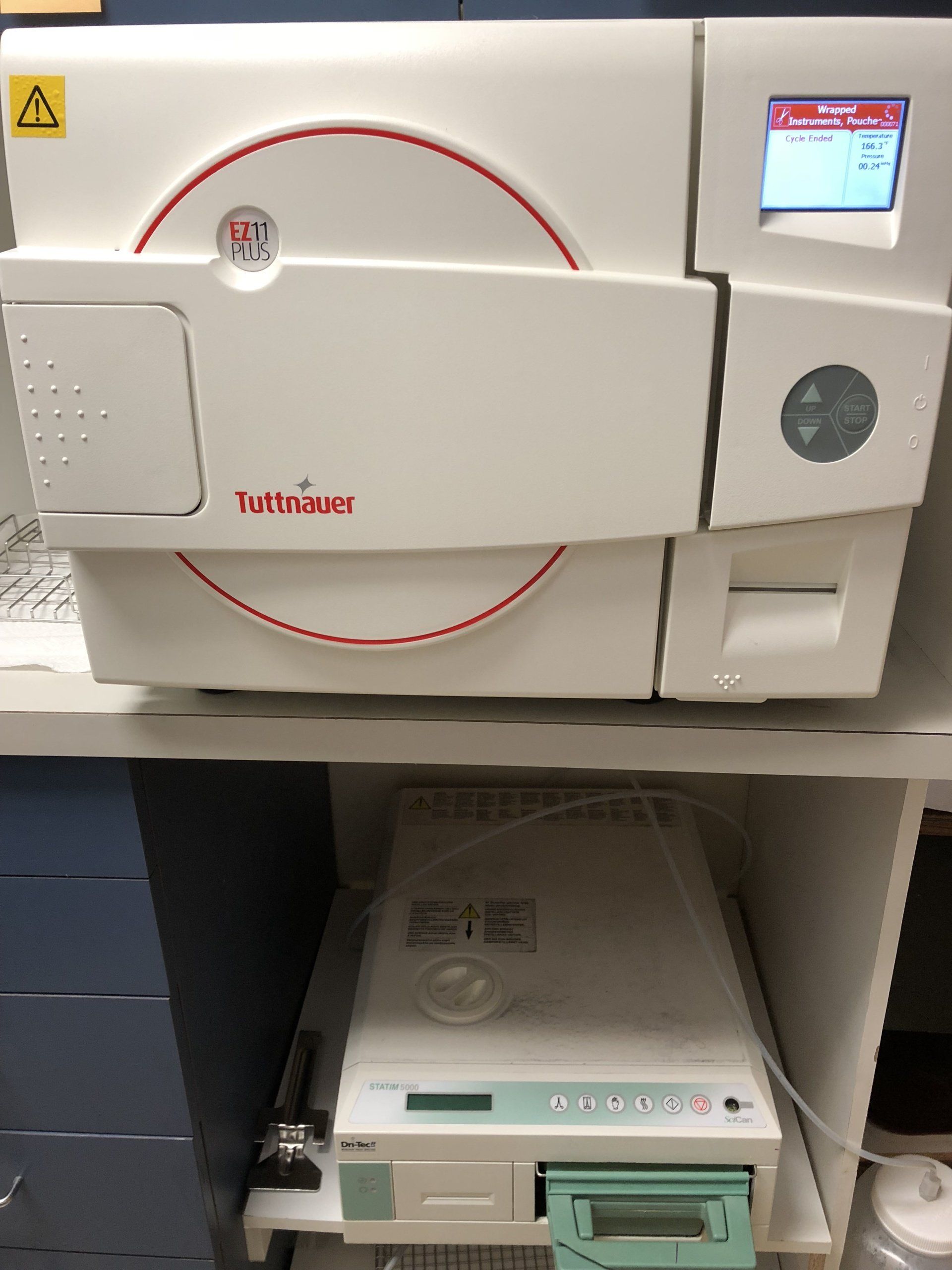A Tuttnauer autoclave sits on a shelf above a water distiller in a clinical setting.