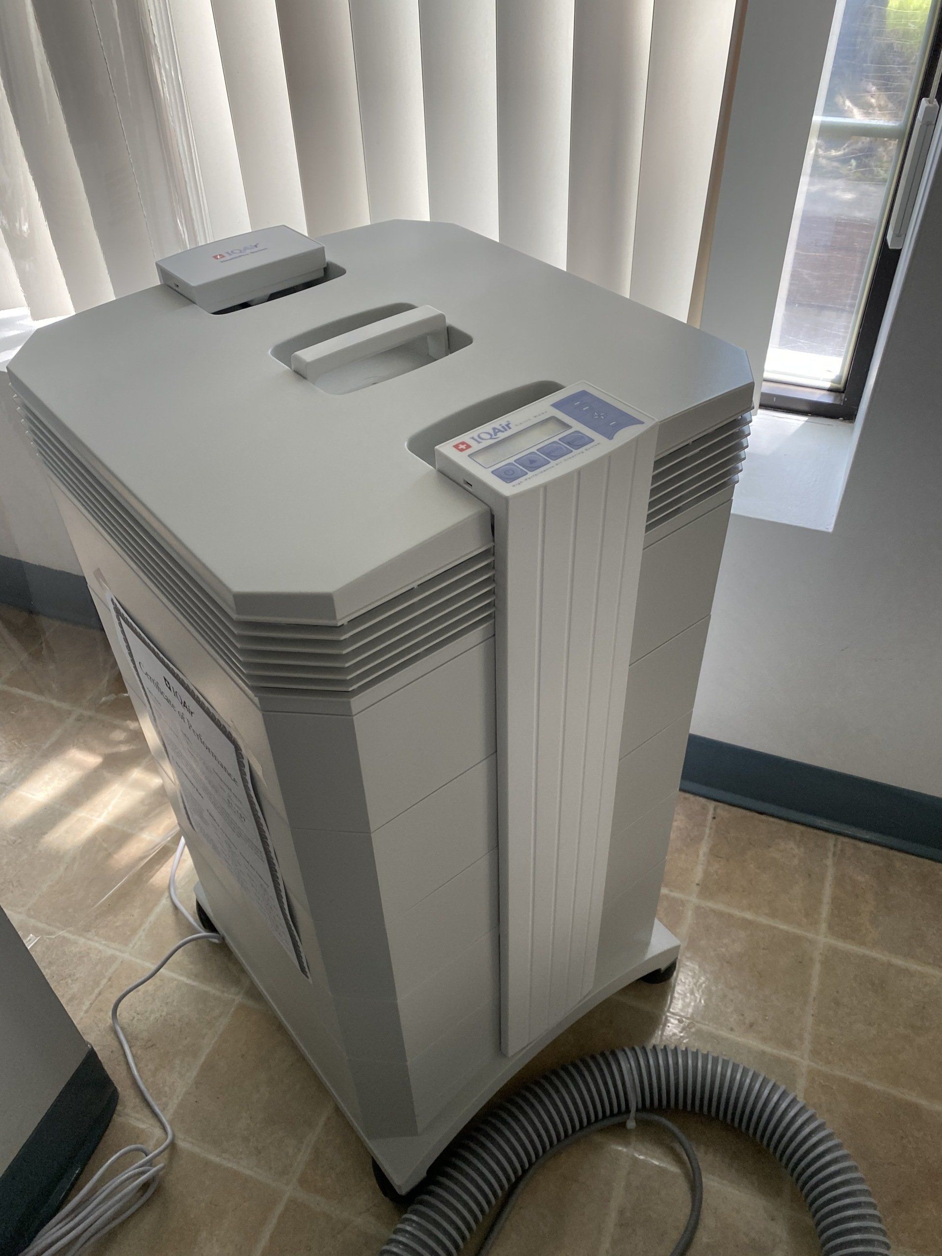 A light gray portable air purifier with a digital control panel, standing on a tile floor near a window with blinds.