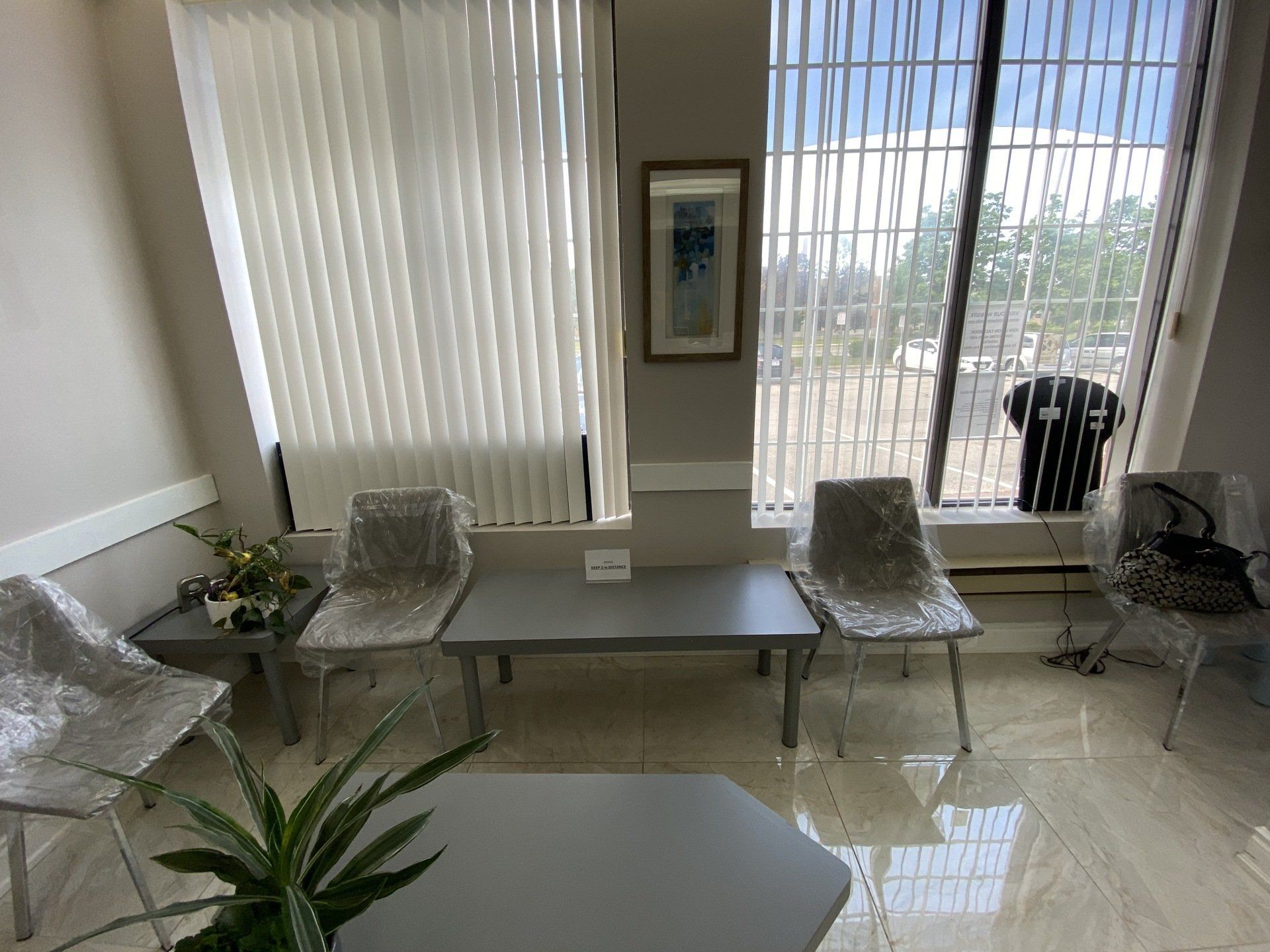 A waiting area with grey chairs, side tables, a plant, wall art, and large windows with vertical blinds.