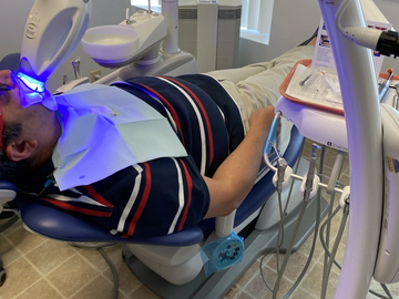 A person reclines in a dental chair during a professional teeth whitening treatment using a blue light lamp.