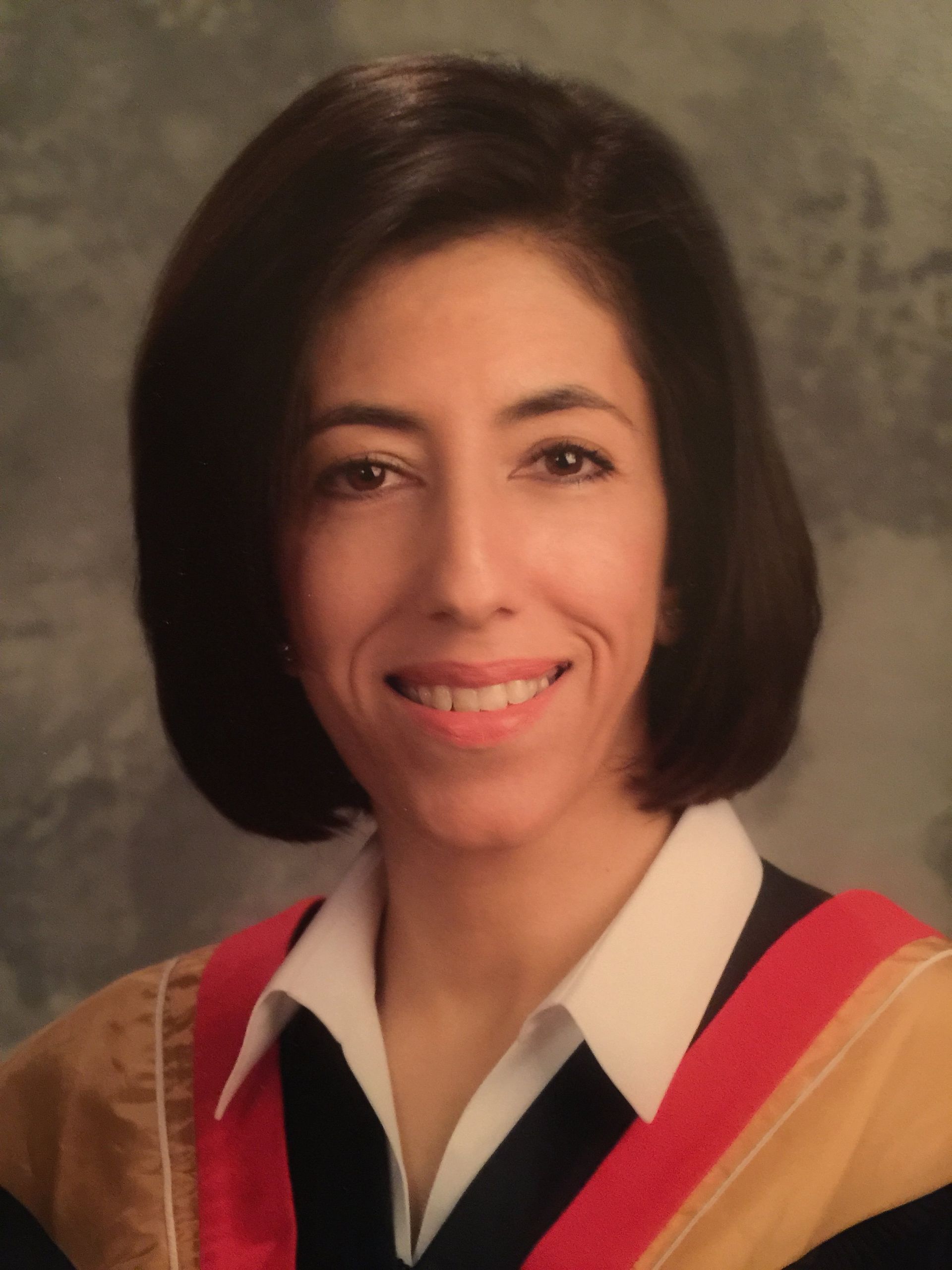 A person with dark hair wearing an academic gown and a collared shirt, smiling against a mottled grey background.