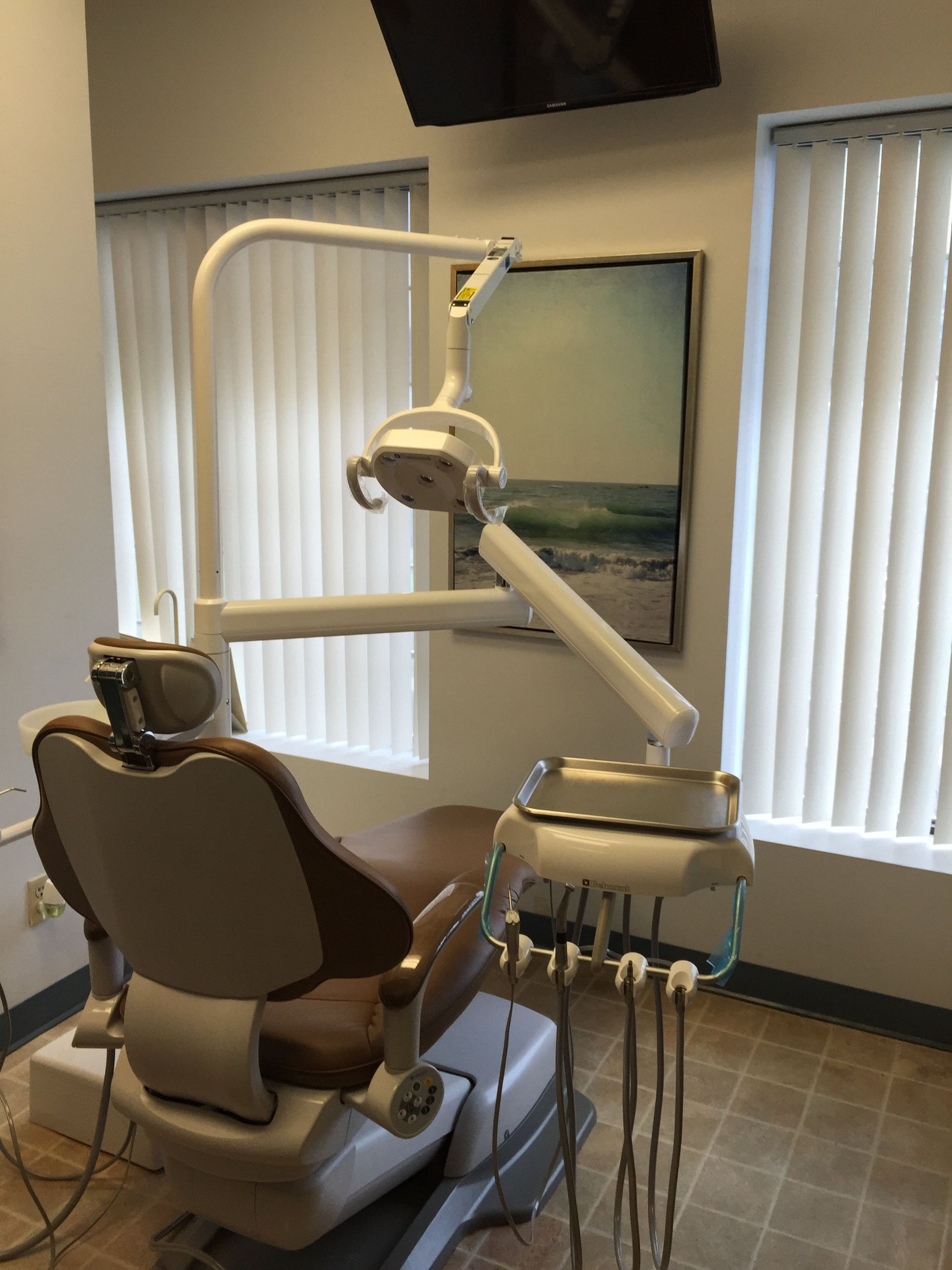 A brown dental chair with an attached light and instrument tray inside a room with vertical blinds and wall art.