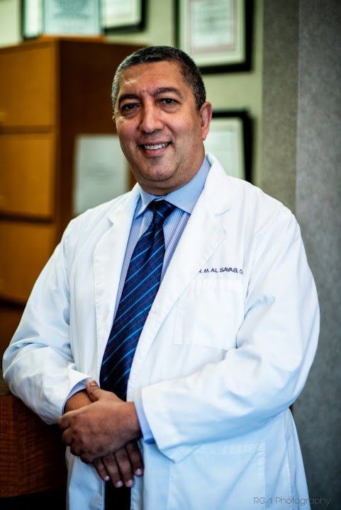 A smiling medical professional in a white coat and blue tie stands with hands clasped in a professional office setting.
