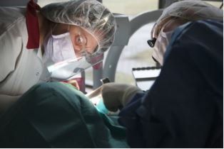 Two healthcare workers in surgical masks, hairnets, and face shields perform a procedure on a patient in a sterile setting.