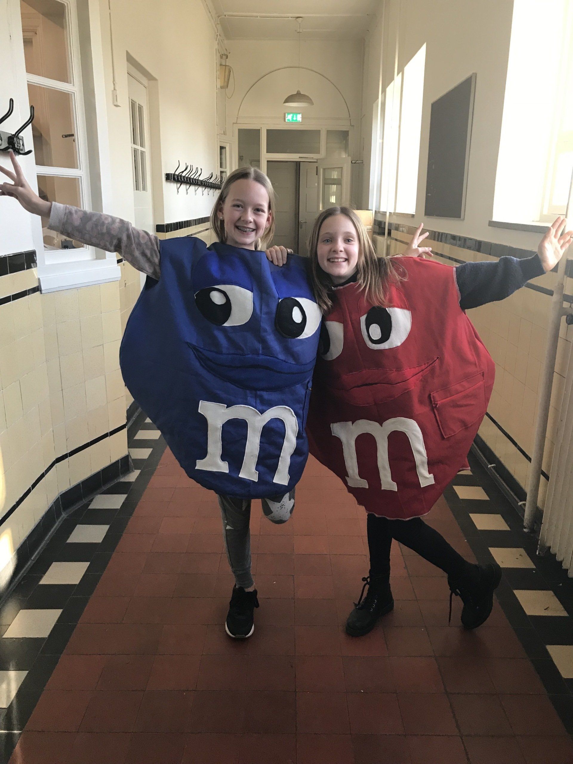 Twee kinderen in M&M-kostuums poseren in een gang. De ene in een blauw, de andere in een rood, met uitgestrekte armen.