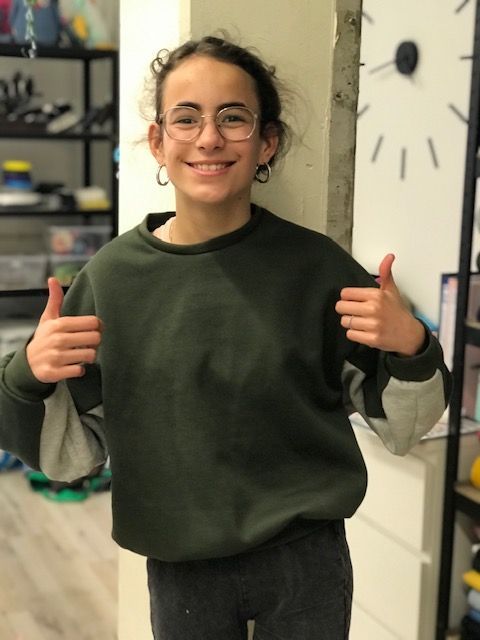 Meisje met bril, duim omhoog, draagt ​​een groen-grijze sweater.