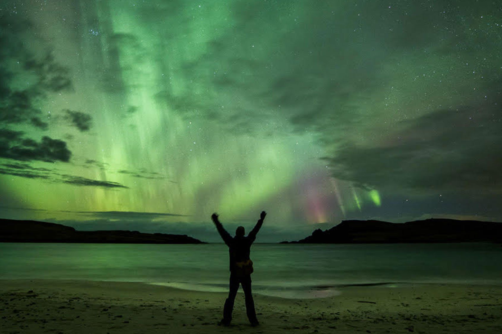 A person is standing on a beach under the aurora borealis.