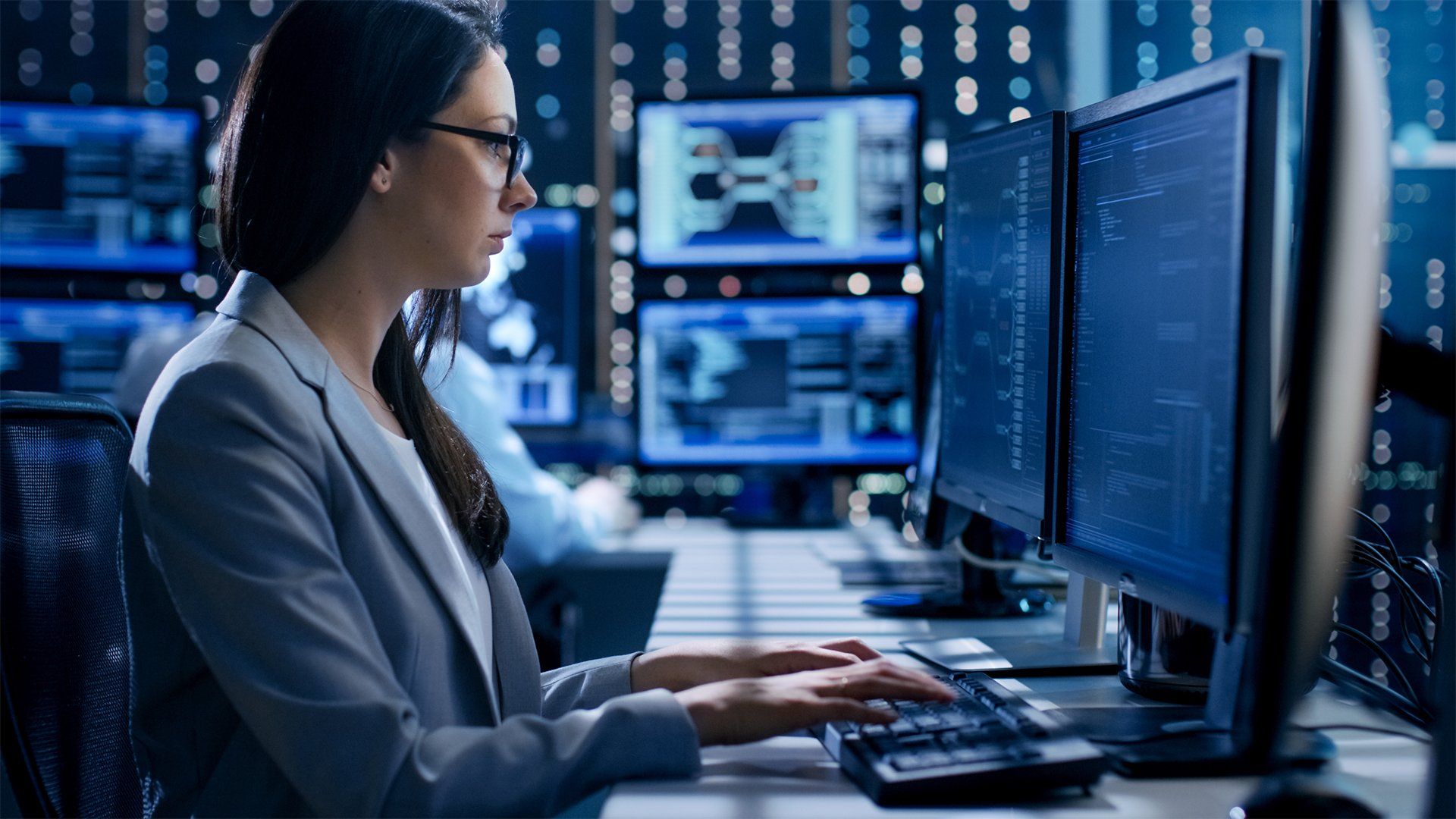 Woman working at a dual-monitor computer in a blue-lit server room