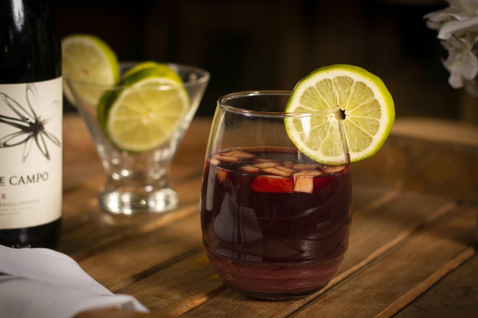 Sangria in a glass with lime garnish, bottle of wine, and sliced limes on a wooden tray.