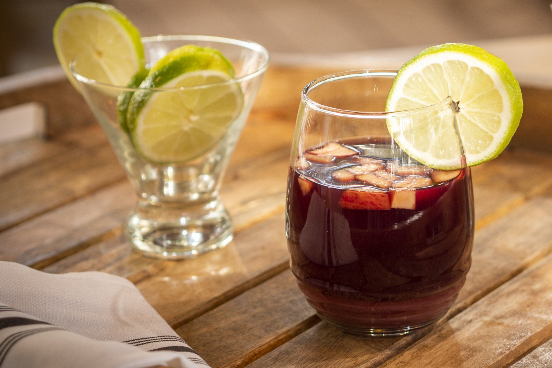 Sangria in a glass with a lime slice, next to a dish of lime slices on a wooden tray.