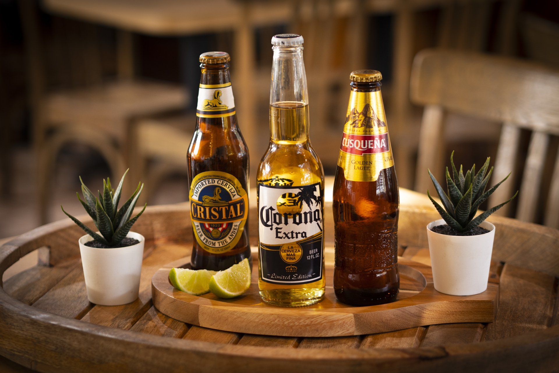 Three beer bottles with limes and small plants on a wooden tray, inside a restaurant.
