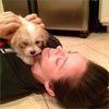 Person lying down, being nuzzled by a small, tan and white dog indoors.
