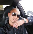 Man in sunglasses, sitting in a car, resting his chin, wearing a black dress shirt and watch.