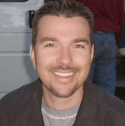 Smiling man with short brown hair, wearing a gray shirt and dark jacket, outside.