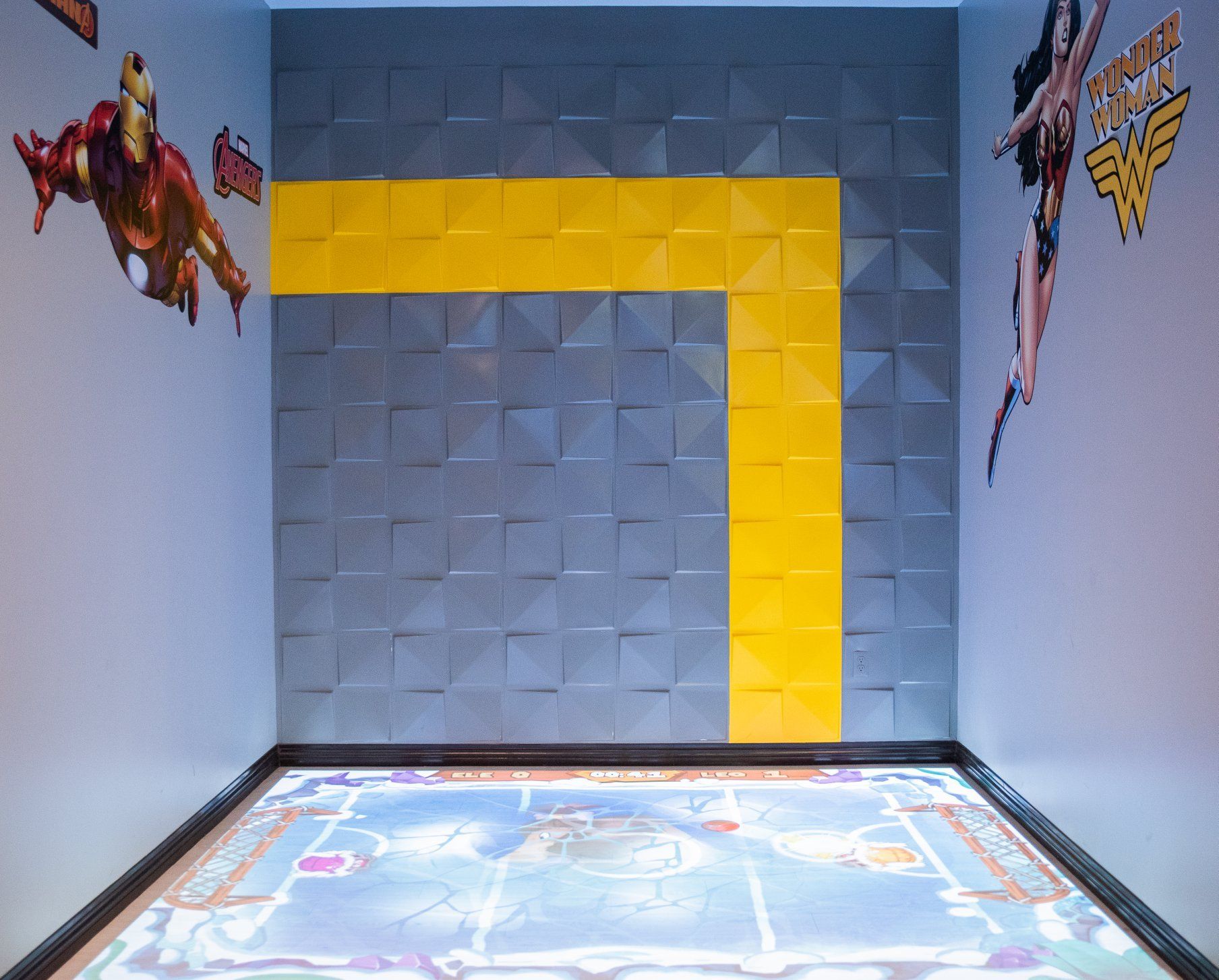Empty room with gray and yellow padded walls and a colorful floor graphic, with superhero decals on the sides
