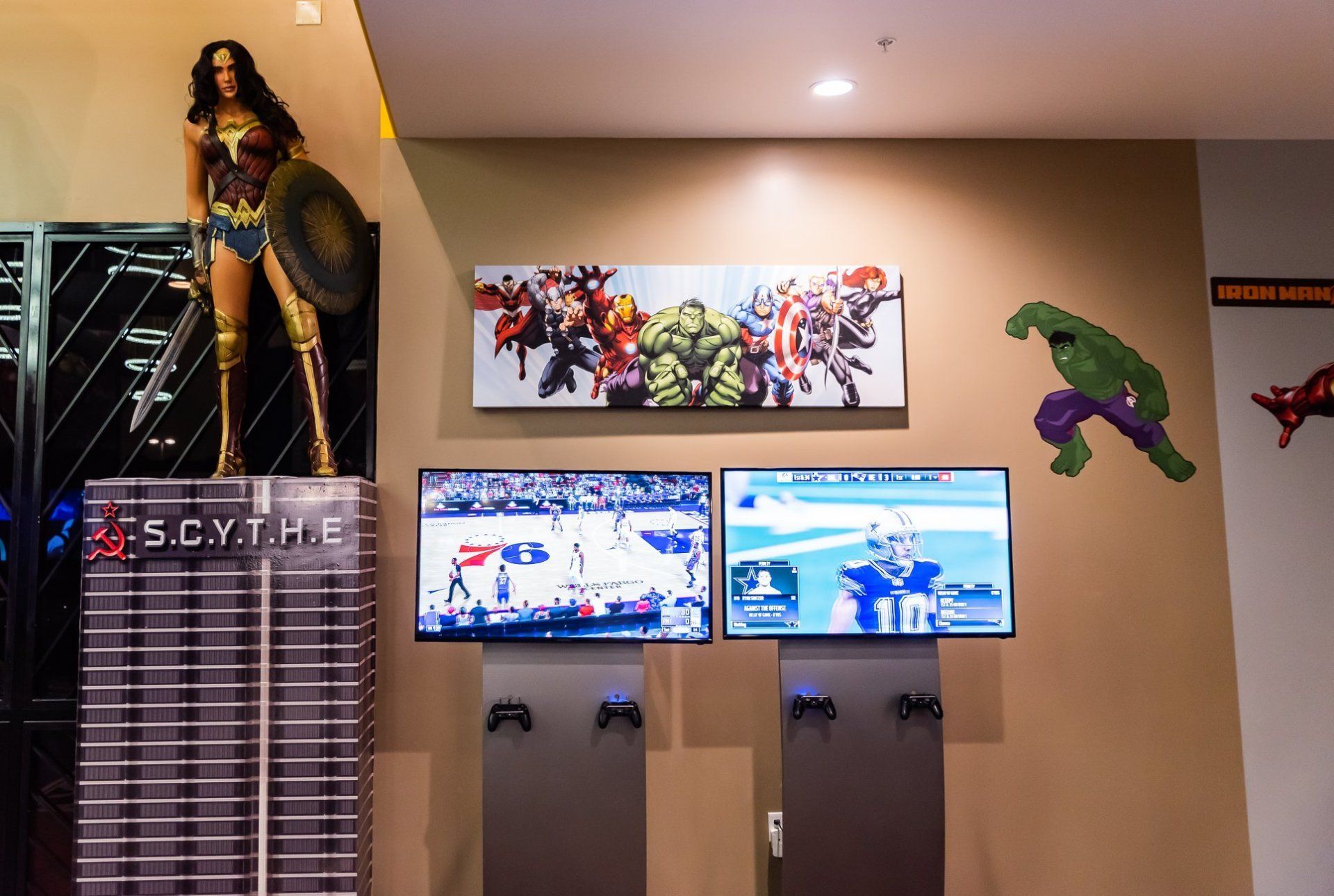 Home arcade room with a wall-mounted TV setup, game posters, and a superhero figure display