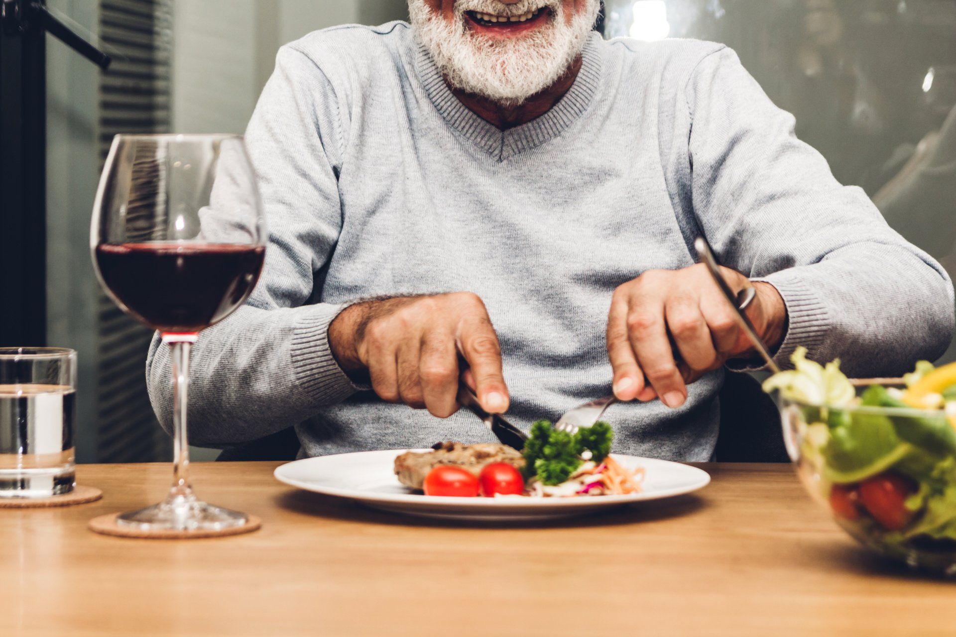 An elderly man is sitting at a table with a plate of food and a glass of wine.