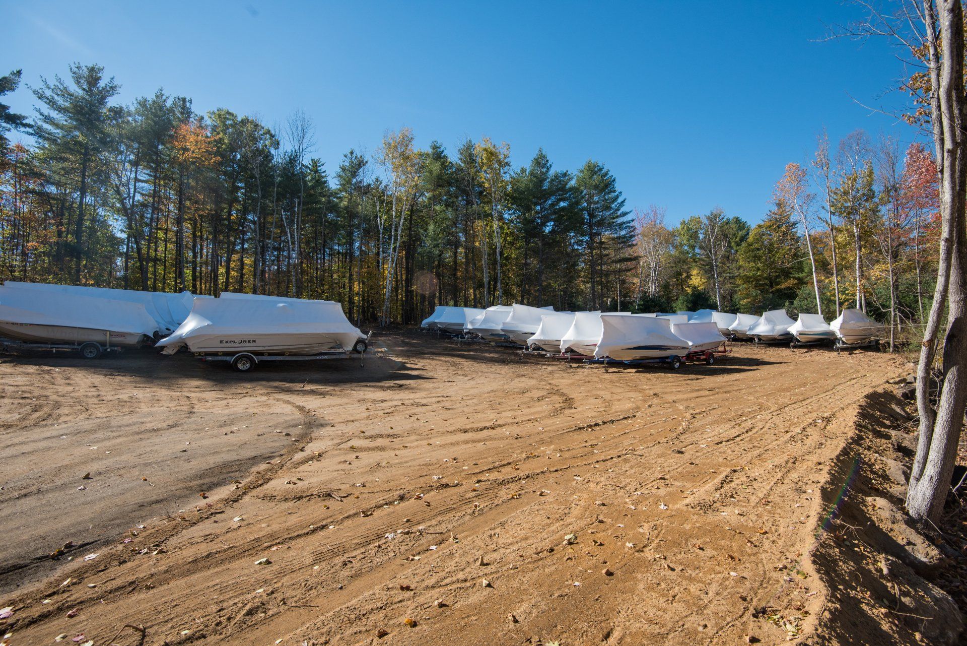 A row of boats covered in plastic are parked in a dirt lot.