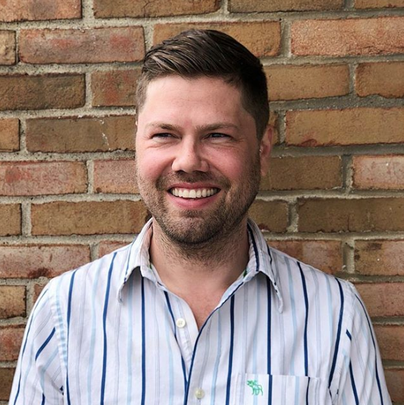 Man with a light beard smiles in front of a brick wall. He wears a striped shirt.
