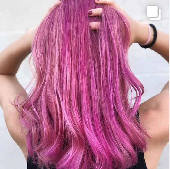 Woman with vibrantly pink, wavy hair, arms raised, black top.