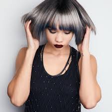 Woman with gray hair, dark lipstick, wearing black tank top, hands up near head, against white background.