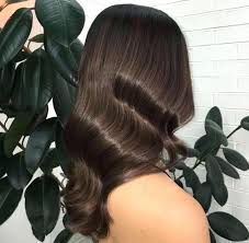 Woman with dark brown, wavy hair styled in retro waves, against a backdrop of green leaves and a white brick wall.