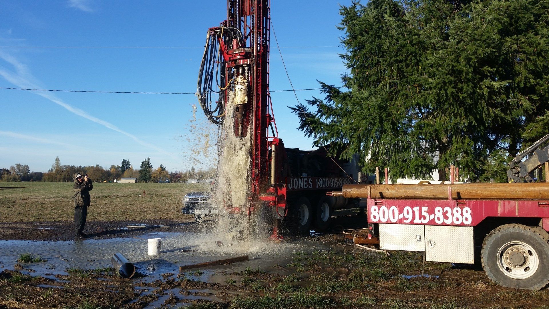 Drilling rig spraying water in field, operator watches, phone number on truck.