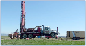 Well drilling rig on a truck, operating in a field on a sunny day.