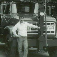 Man in white shirt and jeans standing next to a large black truck, hand on hip.