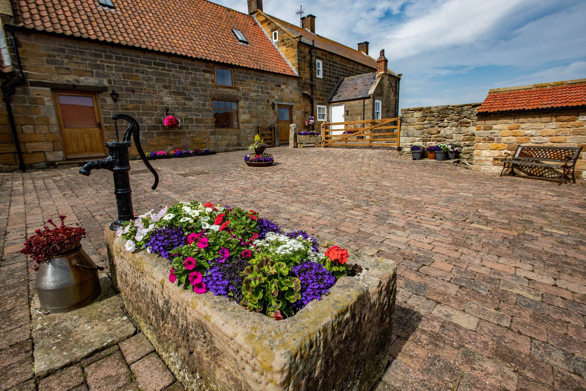 Courtyard with stone buildings, a flower-filled planter, water pump, and brick paving on a sunny day.